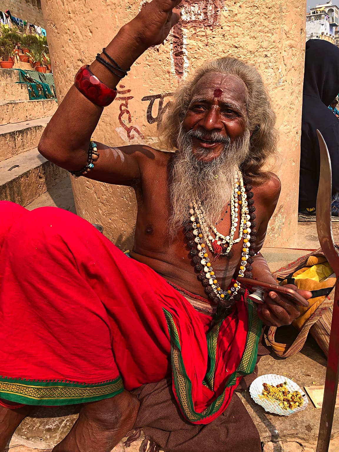 A smiling long-haired sadhu sits in red cloth with one hand raised above the frame in Varanasi, 2019