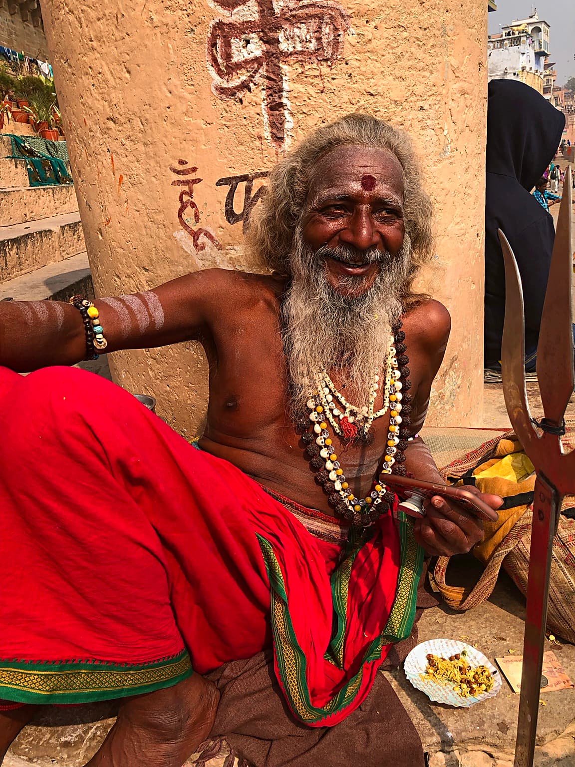 A smiling long-haired sadhu sits in vivid red cloth on the ghats of Varanasi, 2019
