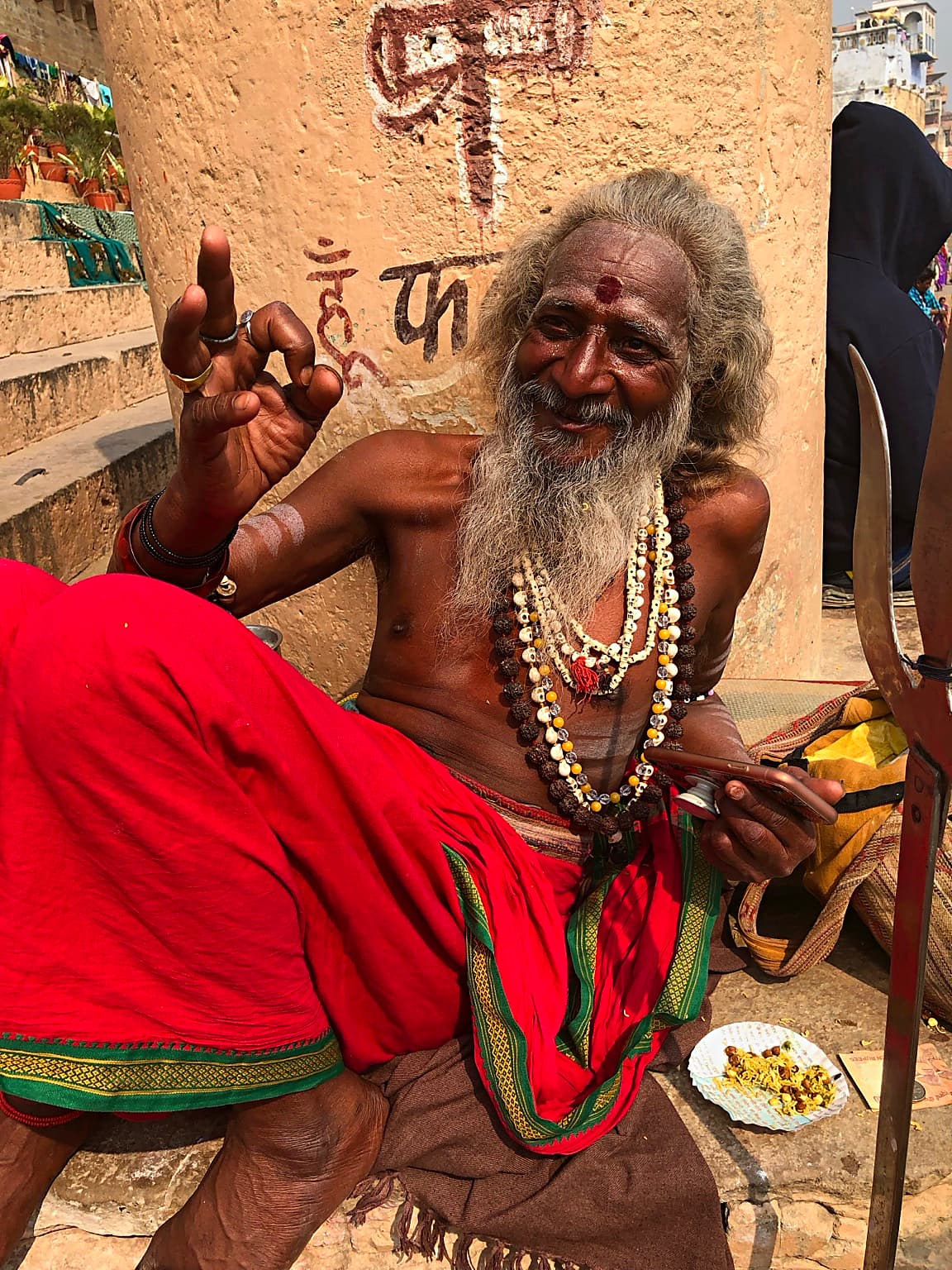 A long-haired sadhu raises one hand while seated in red cloth on the ghats of Varanasi, 2019