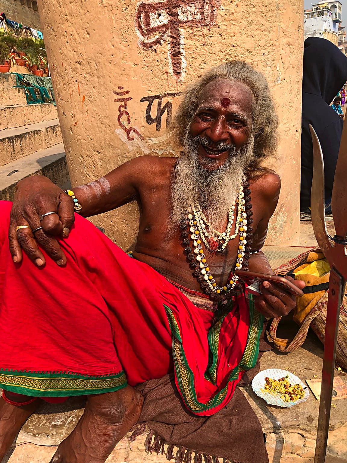 A smiling long-haired sadhu looks toward the camera while seated on the ghats of Varanasi, 2019