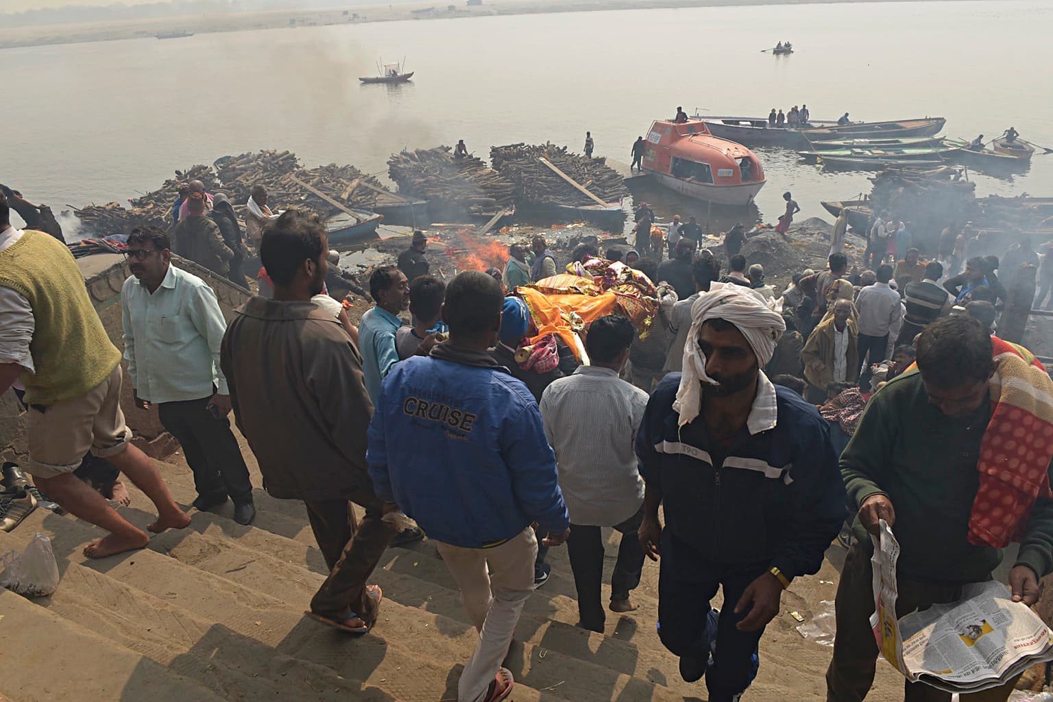 People gather among stacked firewood, smoke and boats at the cremation ghats of Varanasi, 2019