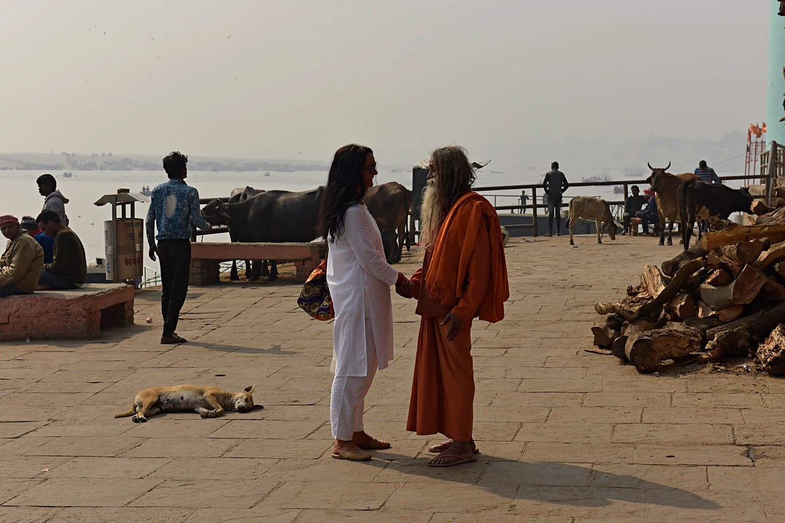 Two men greet one another beside cattle, a resting dog, stacked firewood and the Ganges in Varanasi, 2019
