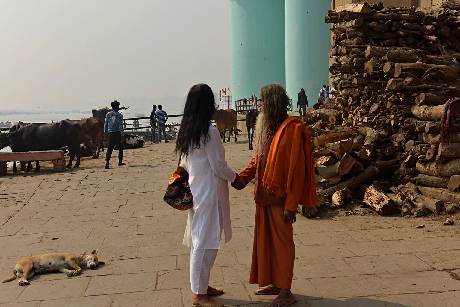 Two men greet one another beside a massive stack of firewood near the Ganges in Varanasi, 2019