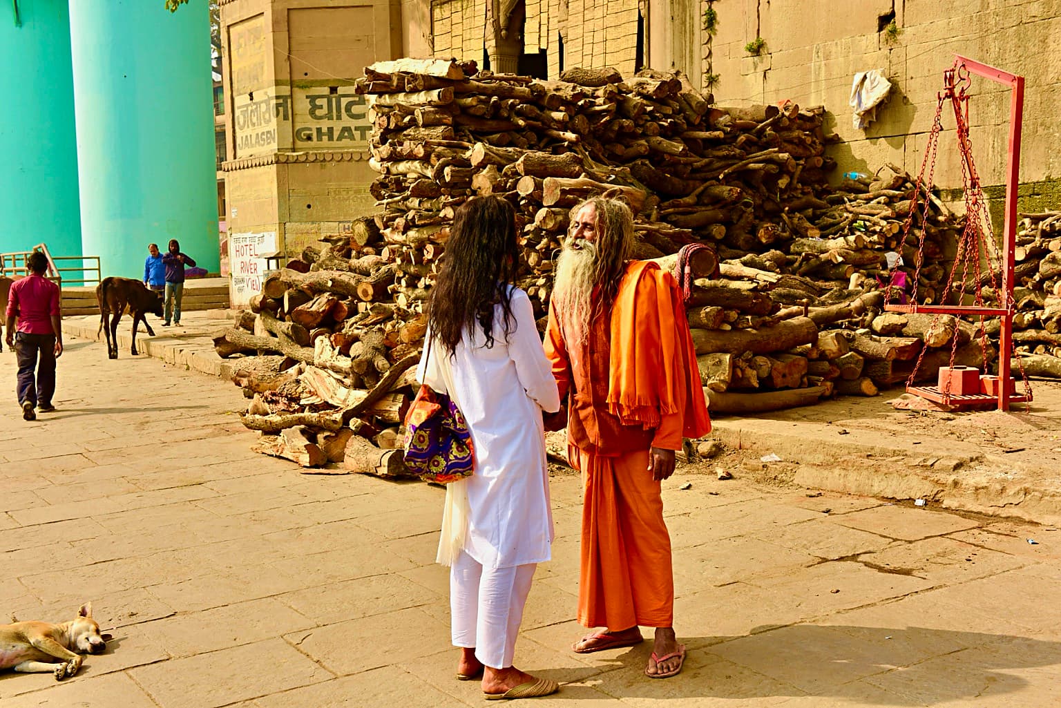 Two people stand talking beside a massive stack of firewood at Jalasen Ghat, with a resting dog nearby, Varanasi, 2019