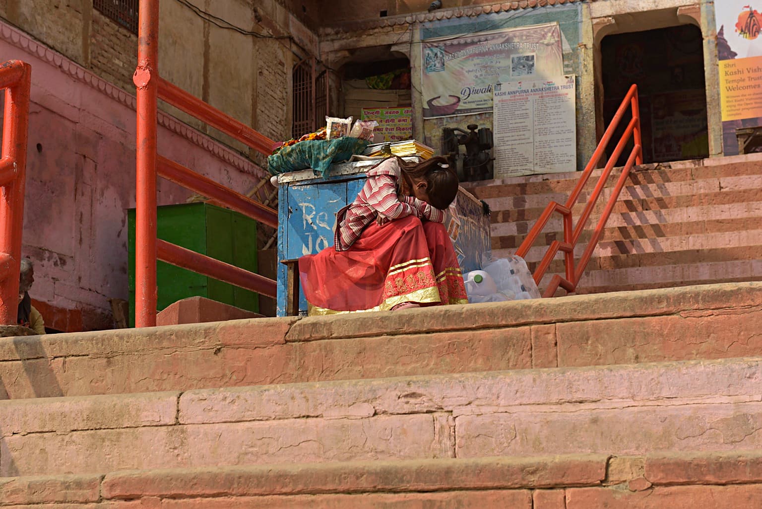 A child sits with head lowered on the ghat steps beside a blue box and red railings in Varanasi, 2019