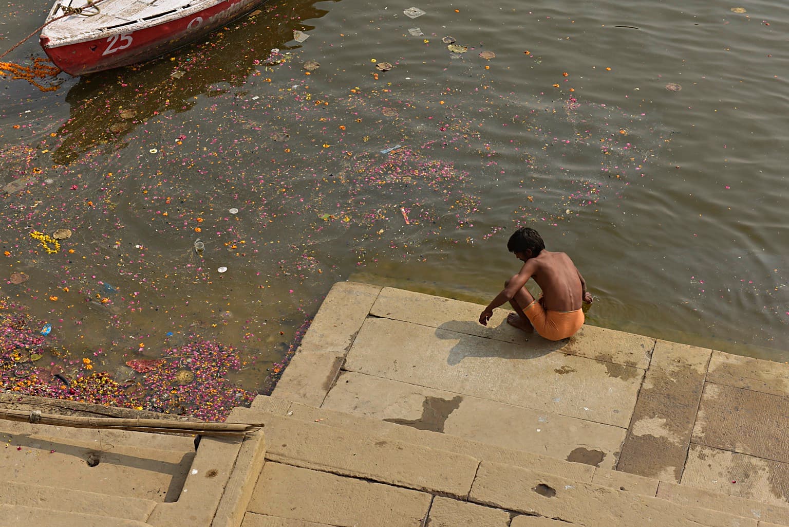 A solitary child sits at the edge of the Ganges amid scattered flower offerings, with part of a boat entering the frame in Varanasi, 2019