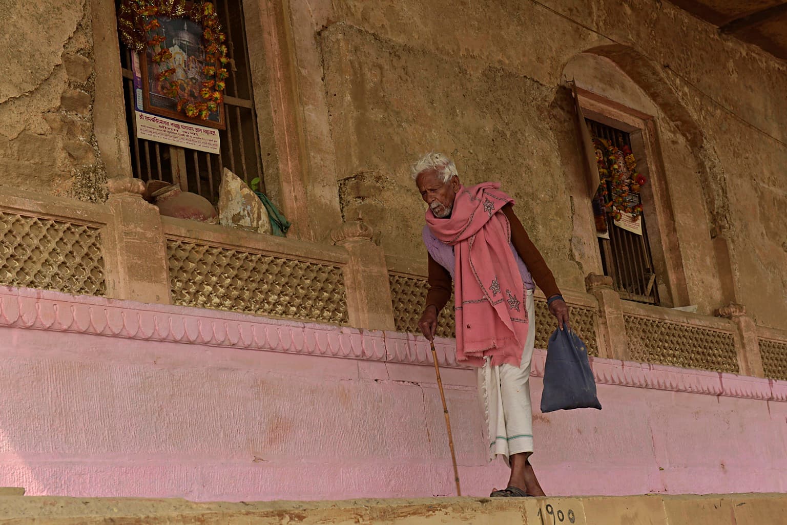 An older man with white hair, a pink shawl and a cane stands before pink riverfront architecture in Varanasi, 2019