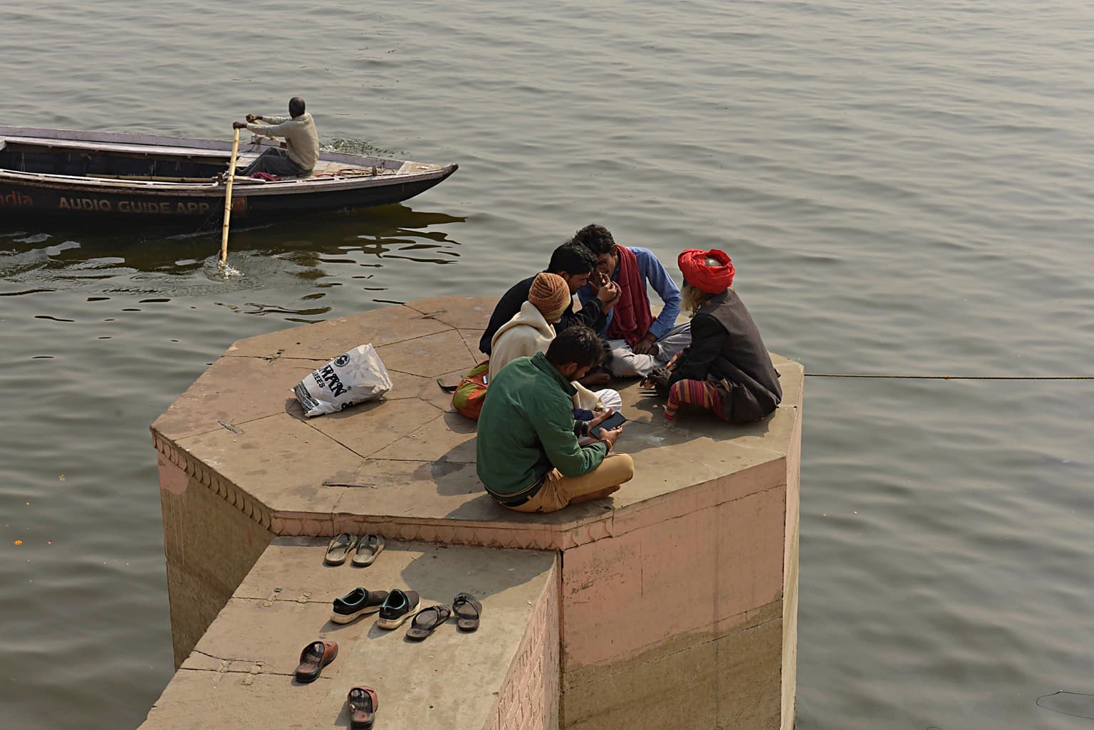 A small group of men gathers on a projecting platform at the Ganges while a rowboat passes nearby in Varanasi, 2019