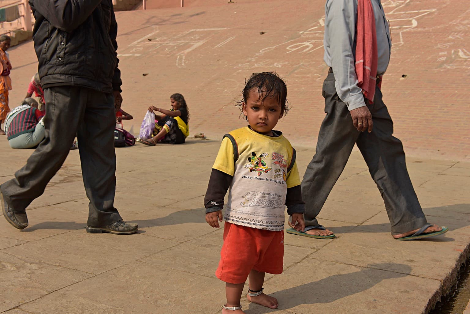 A barefoot young child in a yellow shirt looks directly toward the camera while adults pass across the ghats in Varanasi, 2019