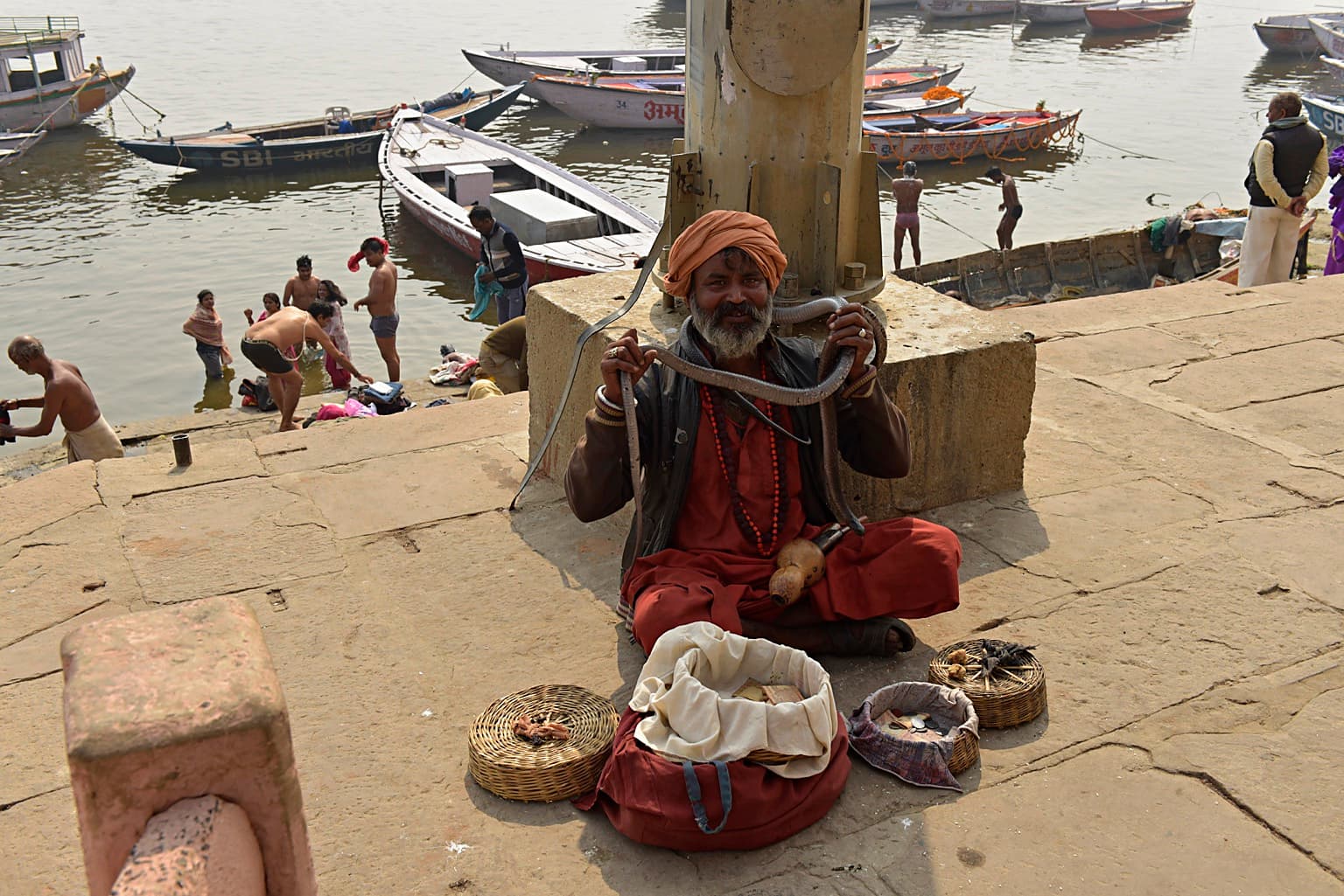 A seated snake charmer holds a snake on the ghats with baskets and the Ganges behind him, Varanasi, 2019