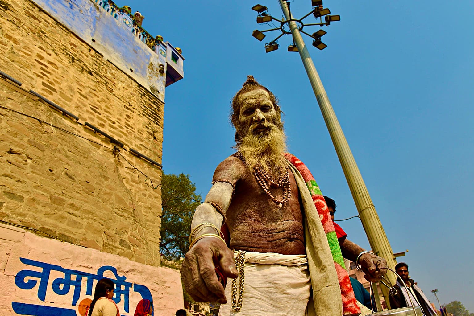 An ash-covered sadhu is photographed from a low angle beneath a clear blue sky on the ghats of Varanasi, 2019