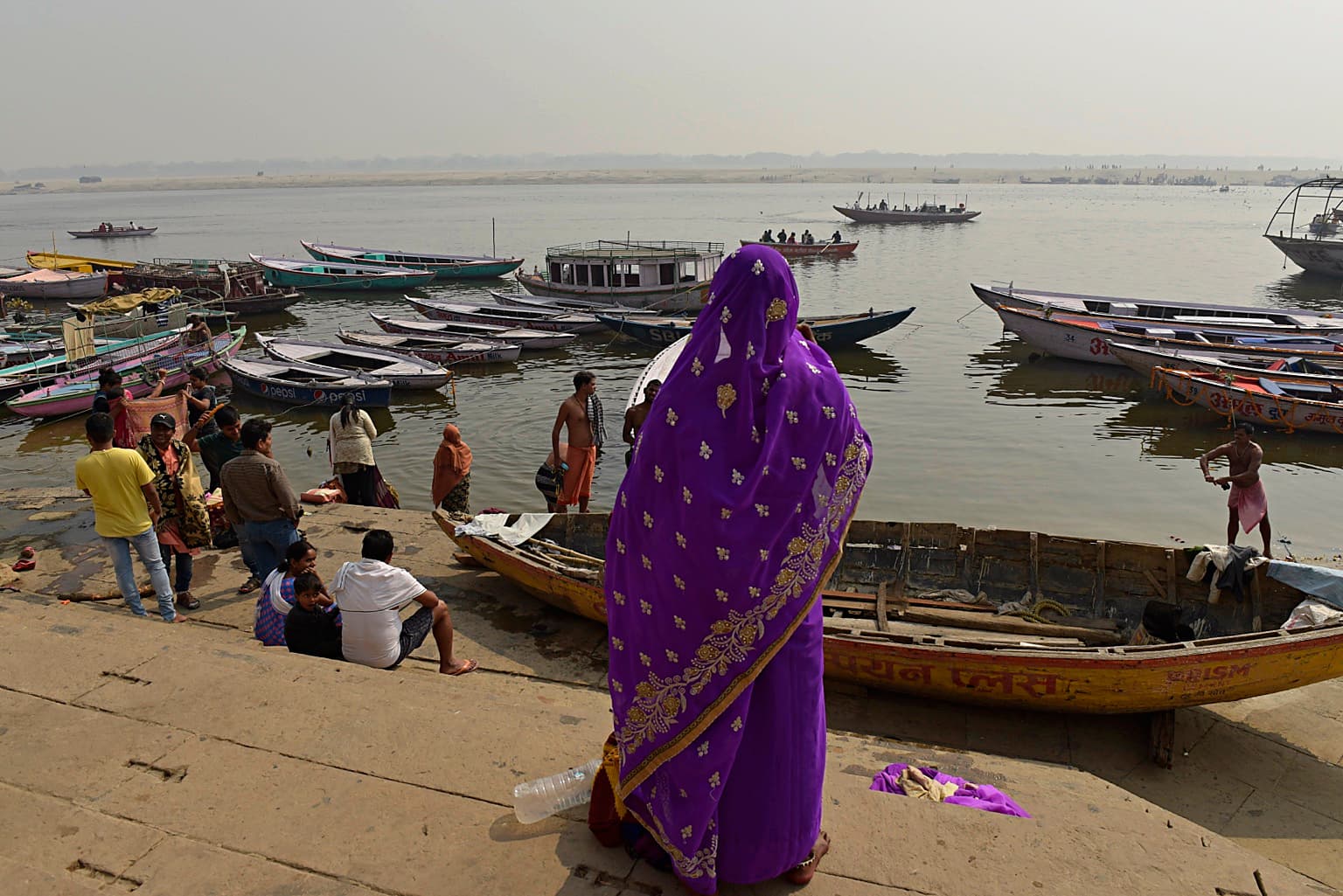 A woman in a vivid purple sari stands on the ghat steps above boats on the Ganges in Varanasi, 2019