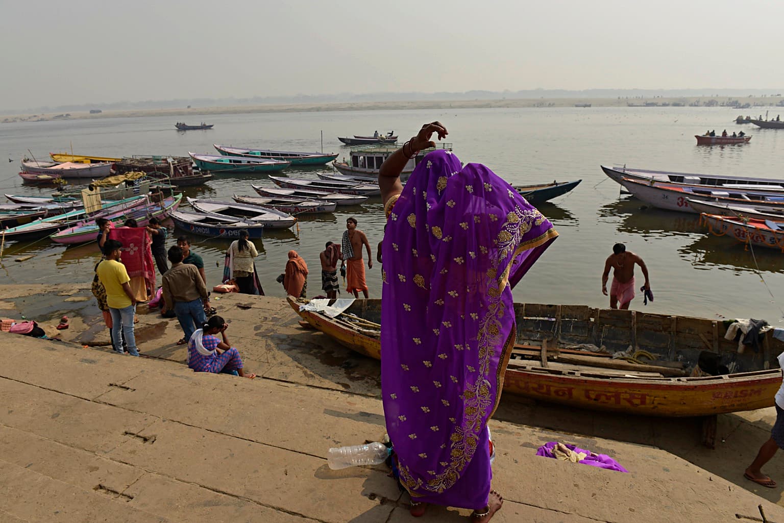 A woman in a purple sari raises one arm while standing above the Ganges and moored boats in Varanasi, 2019