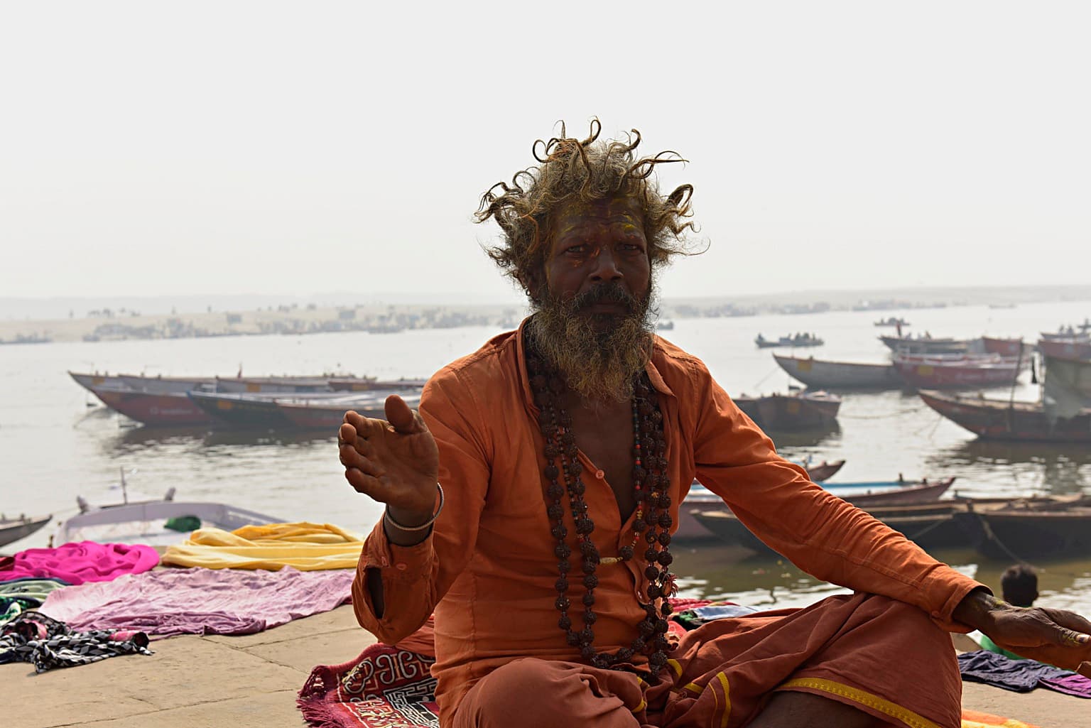 A seated sadhu in orange raises one hand with the Ganges and moored boats behind him, Varanasi, 2019