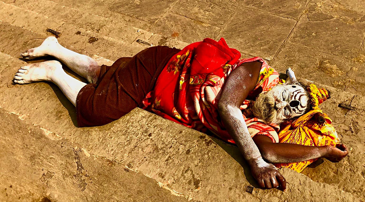 A person with white-painted face and limbs lies resting on the stone ghats beneath red and orange cloth in Varanasi, 2019