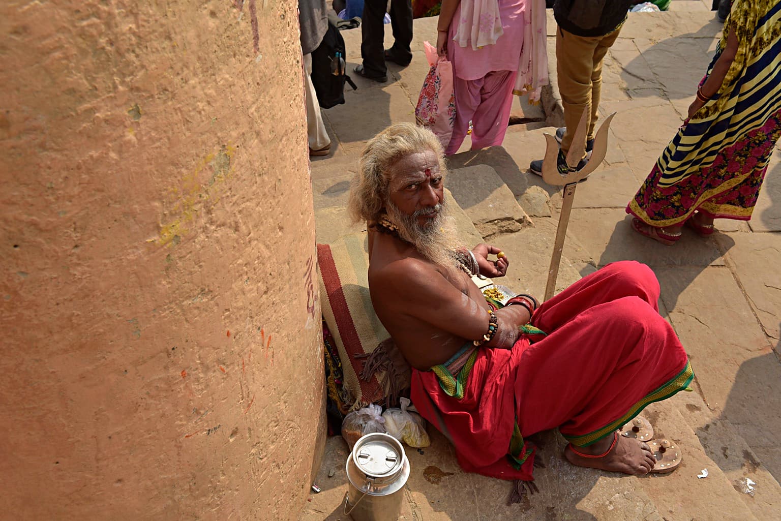 A seated long-haired sadhu in red cloth looks up toward the camera beside his trident in Varanasi, 2019