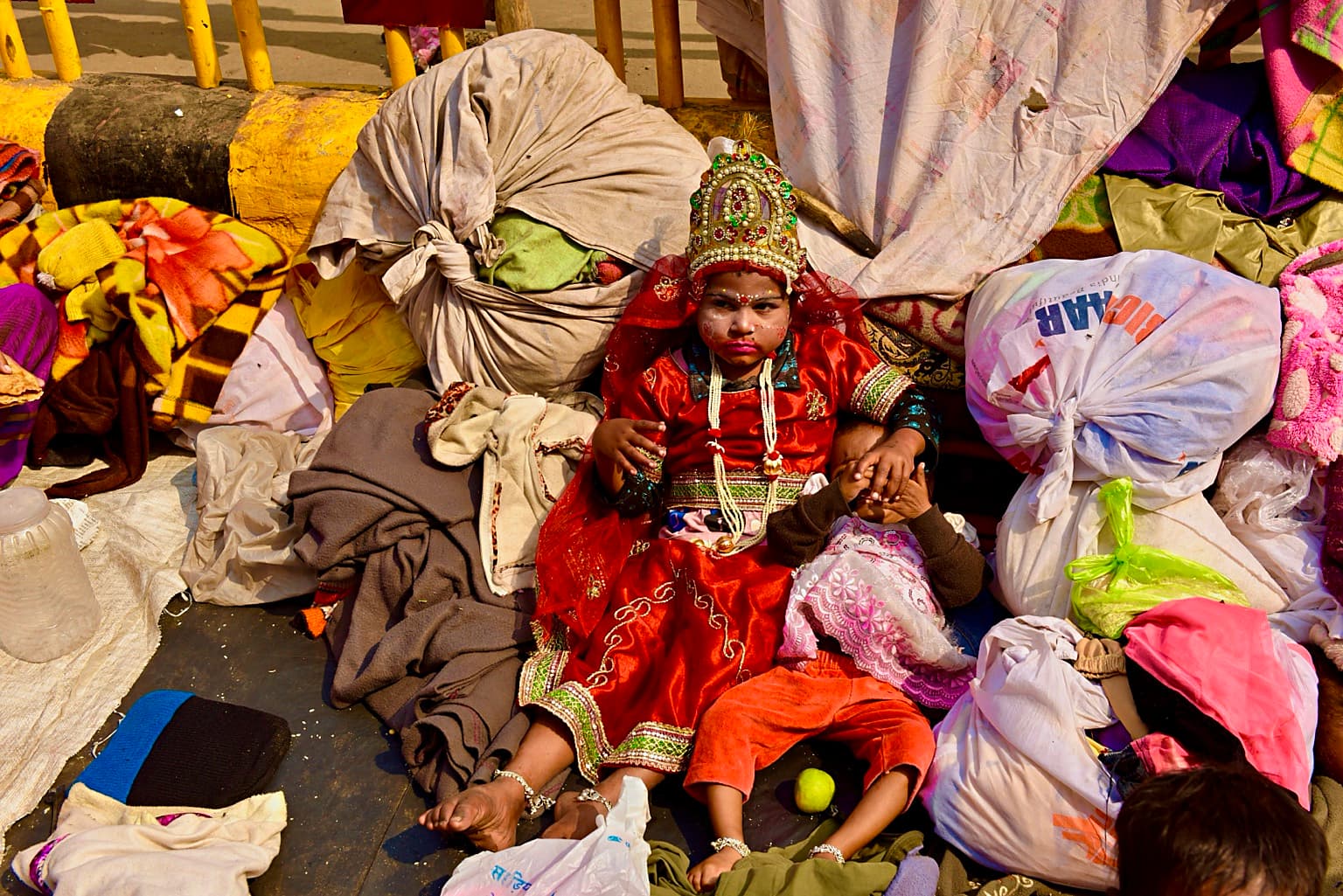 A child in elaborate red ceremonial dress sits among blankets and bundled belongings in Varanasi, 2019