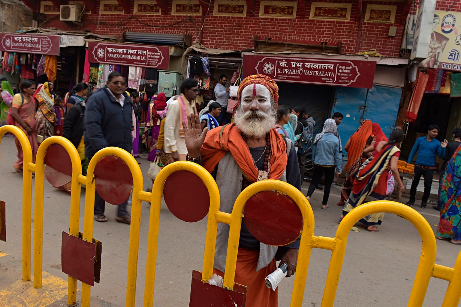 A white-faced sadhu raises one hand behind a bright yellow street barrier in Varanasi, 2019