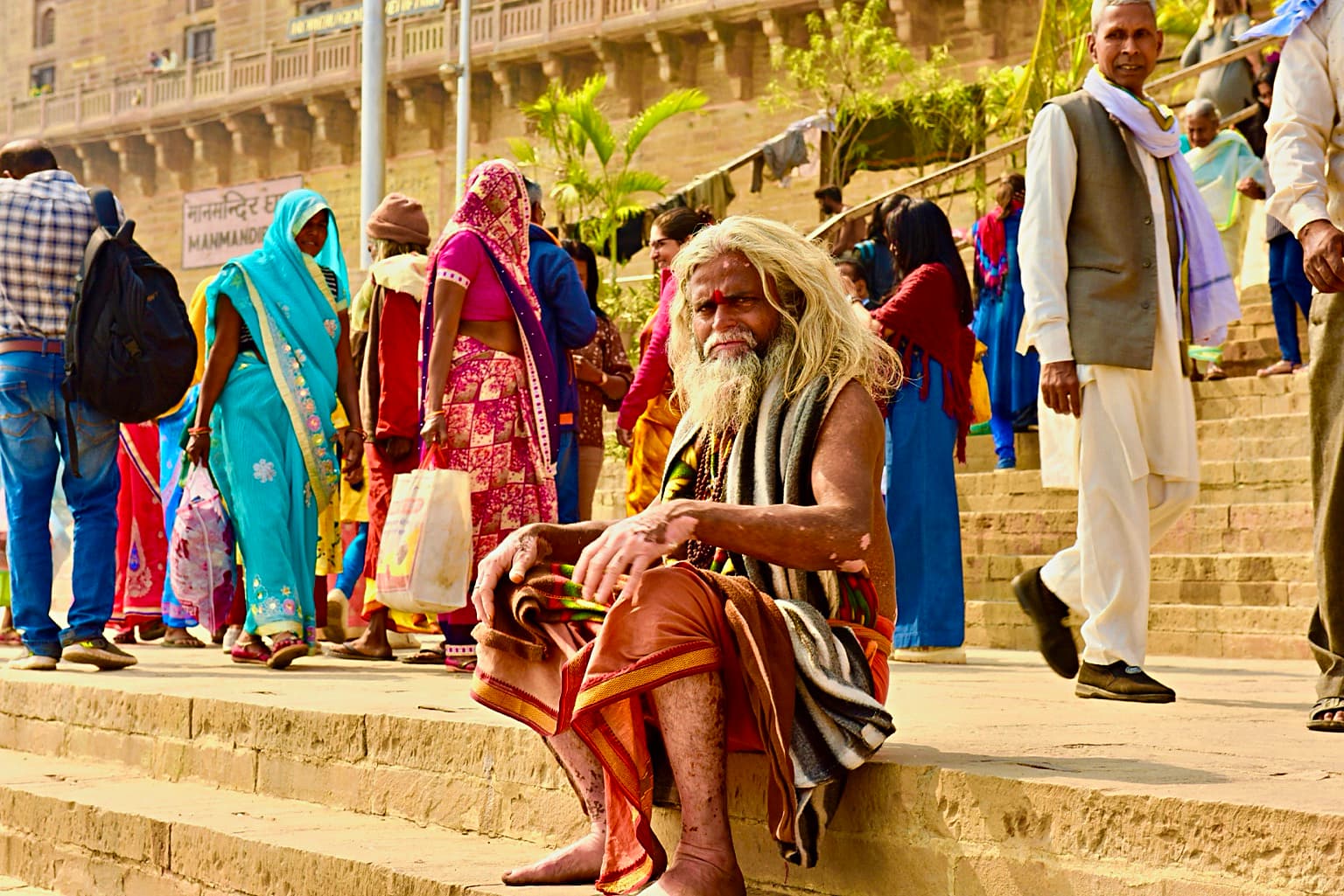 A long-haired sadhu sits on the ghat steps while brightly dressed pedestrians move behind him in Varanasi, 2019