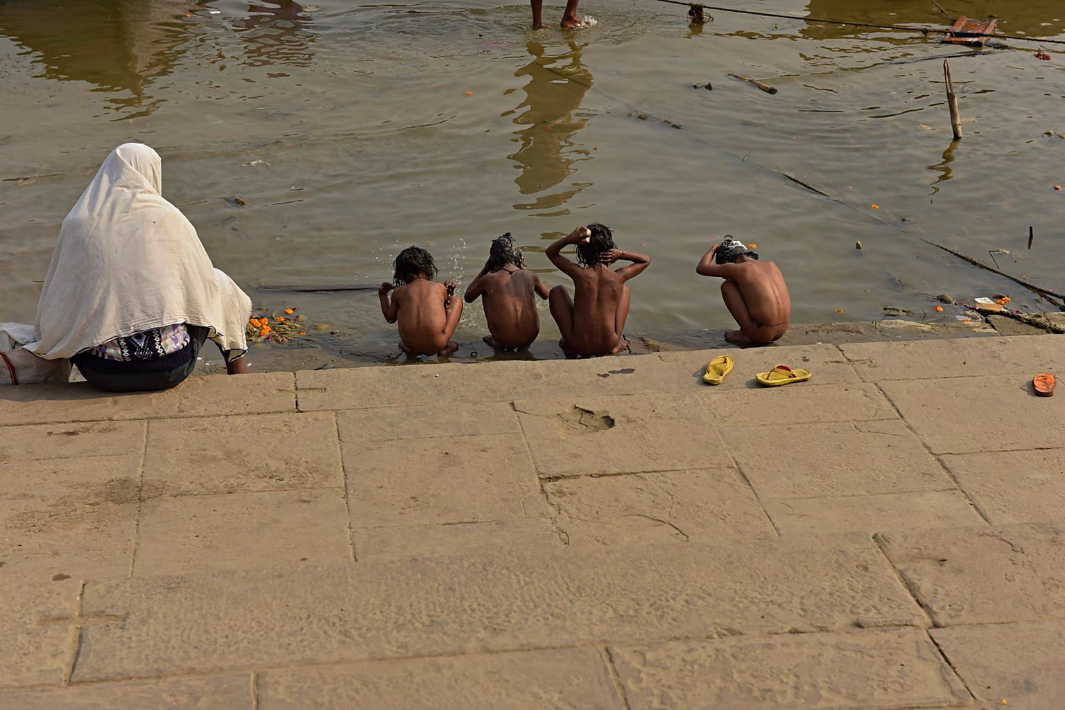 Four children bathe at the edge of the Ganges while an adult sits wrapped in white nearby, Varanasi, 2019