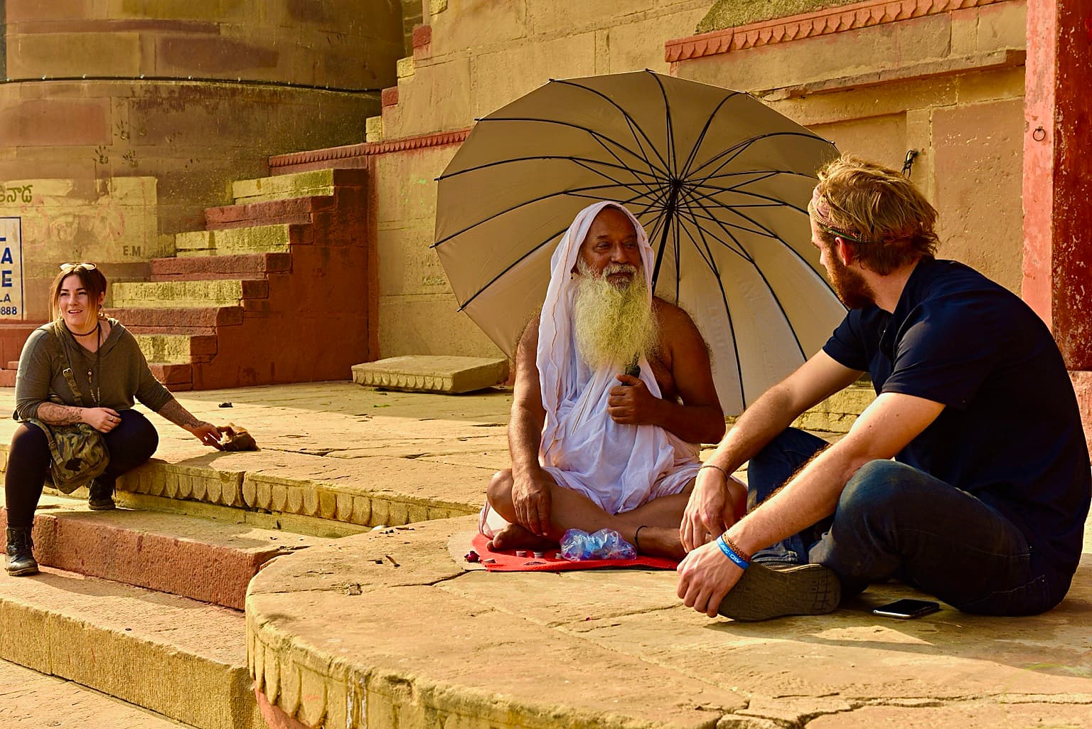 A white-bearded holy man sits beneath a large umbrella speaking with two visitors on the ghats of Varanasi, 2019