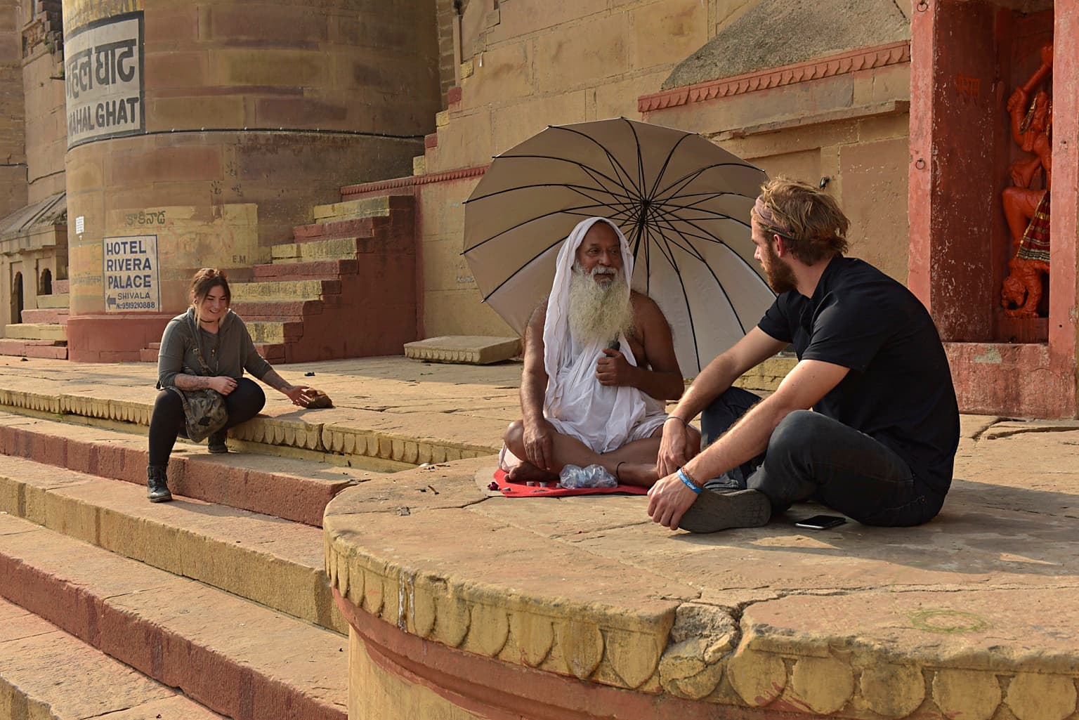 A wider view shows a white-bearded holy man beneath an umbrella with two visitors amid the architecture of the ghats, Varanasi, 2019