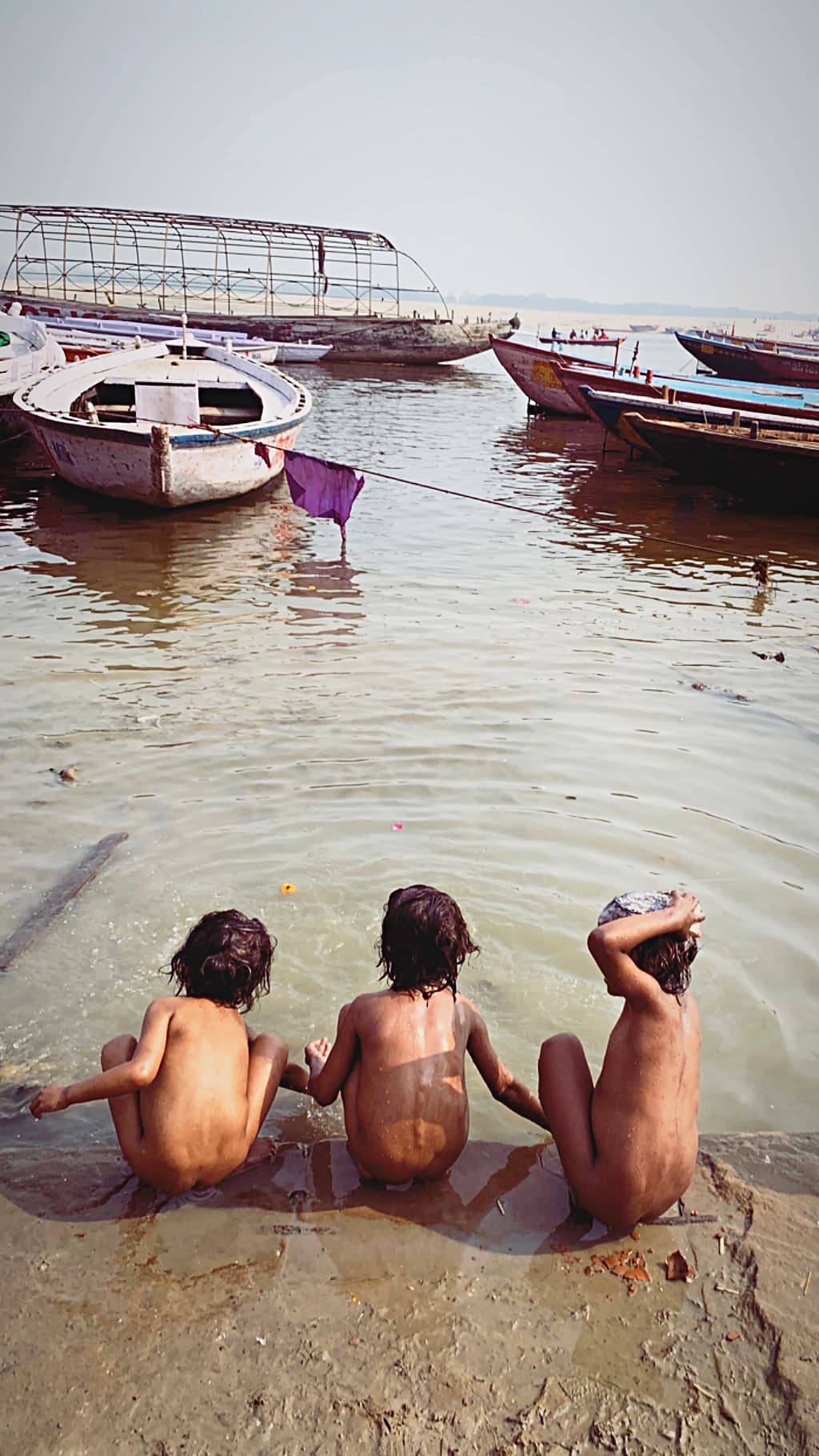Three children bathe at the edge of the Ganges with wooden boats floating beyond them in Varanasi, 2019