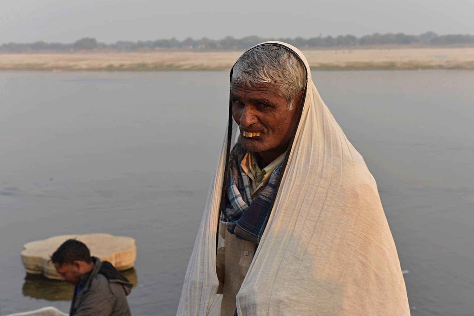 An older man wrapped in a pale shawl stands for a portrait beside the Ganges in Varanasi, 2019