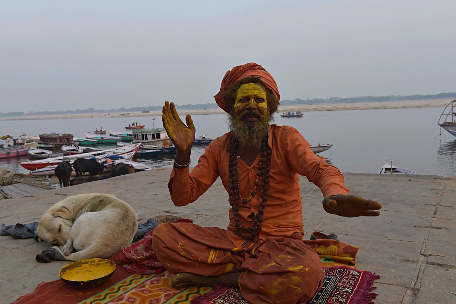 A yellow-pigmented sadhu in orange robes sits beside a sleeping dog and raises one hand above the Ganges in Varanasi, 2019