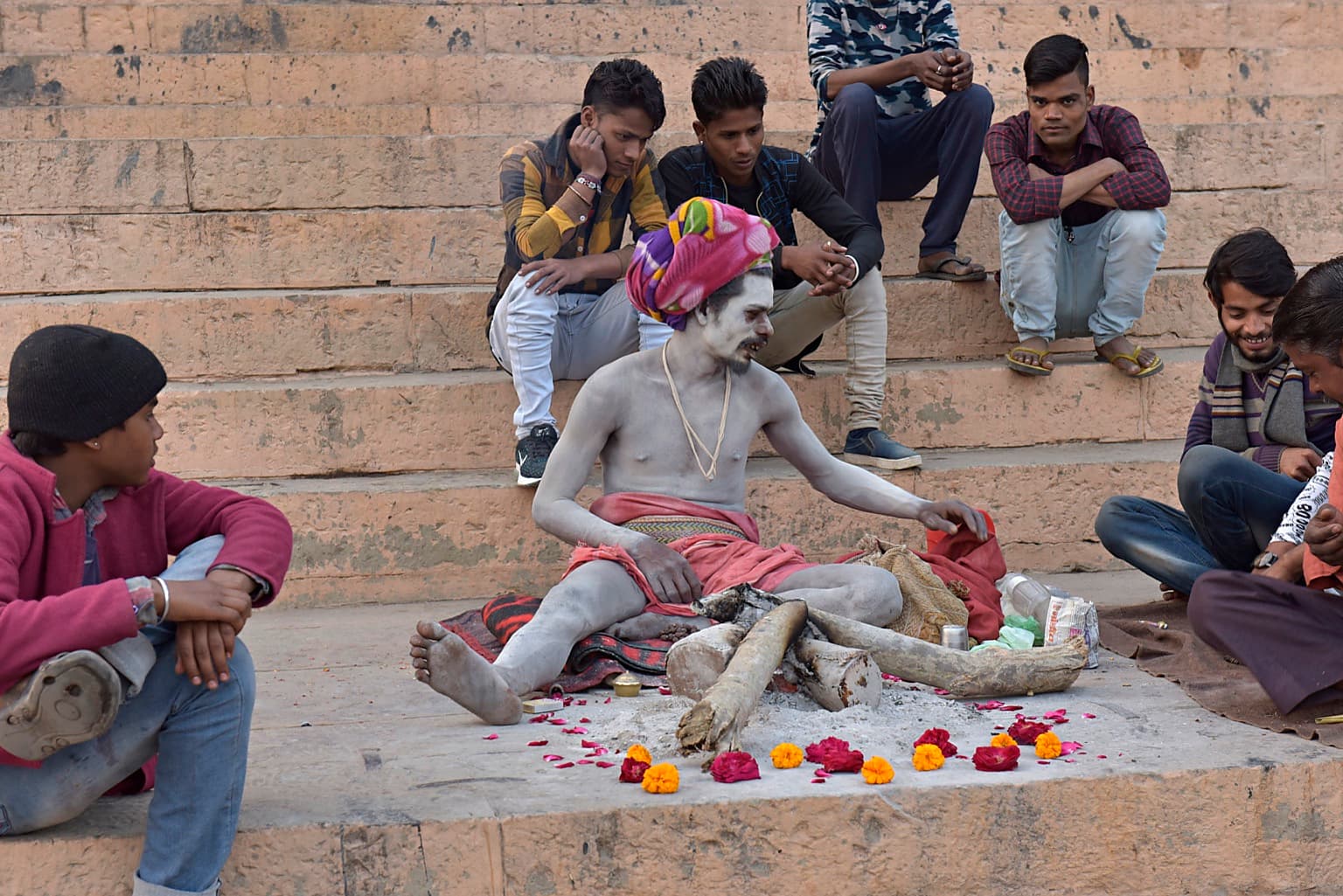 An ash-covered sadhu in a colourful headwrap sits on the stone ghats among onlookers and flower offerings in Varanasi, 2019
