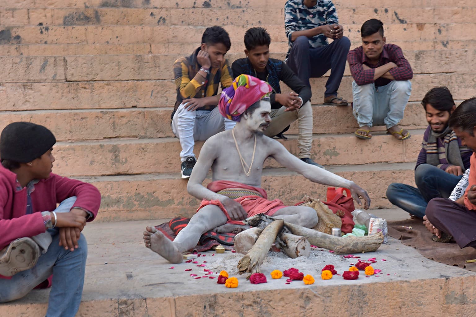An ash-covered sadhu in a colourful headwrap turns toward people seated around him on the ghats of Varanasi, 2019