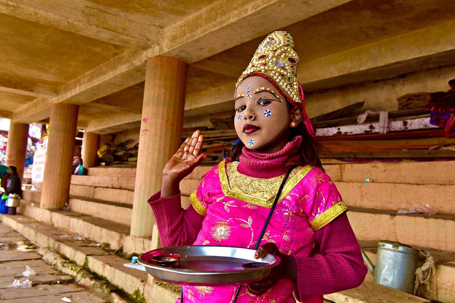 A young girl in bright pink ceremonial dress and a jeweled headpiece raises one hand while holding a metal tray in Varanasi, 2019