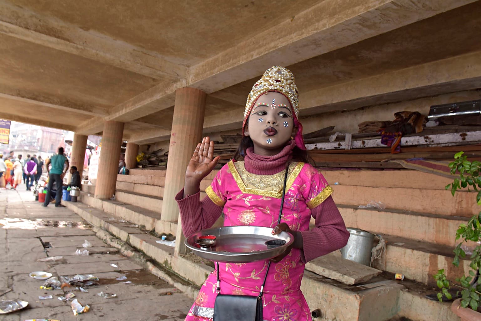 A young girl in bright pink ceremonial dress raises one hand and looks to the side while holding a shallow metal tray in Varanasi, 2019