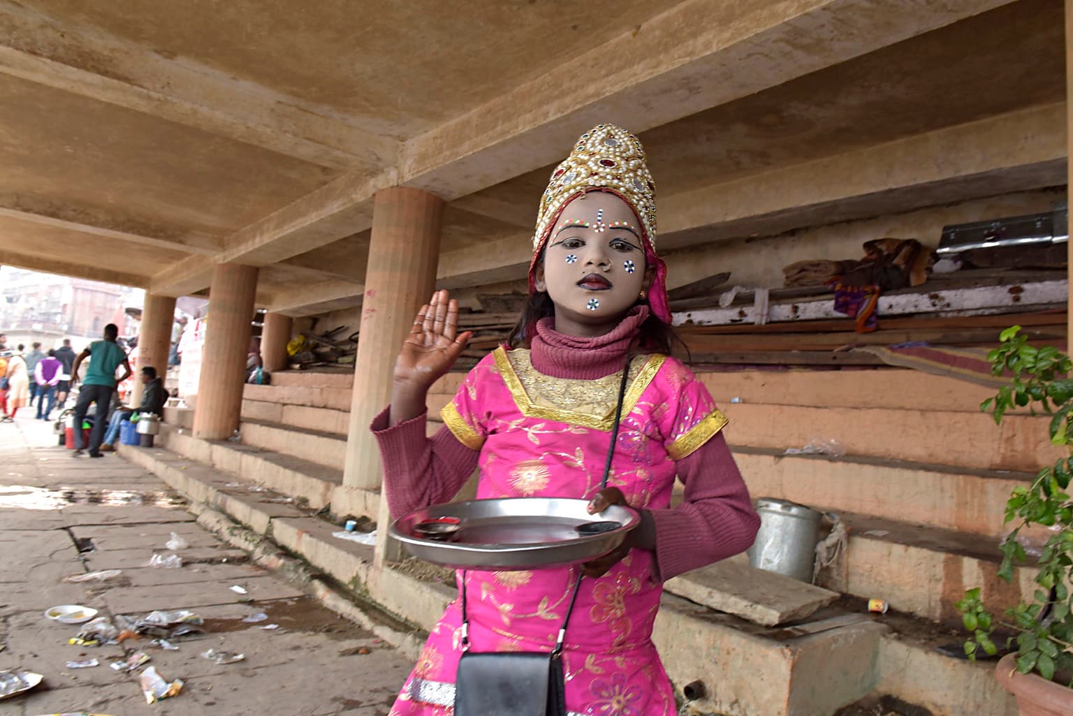 A young girl in bright pink ceremonial dress faces the camera while holding a metal tray beneath the ghats in Varanasi, 2019