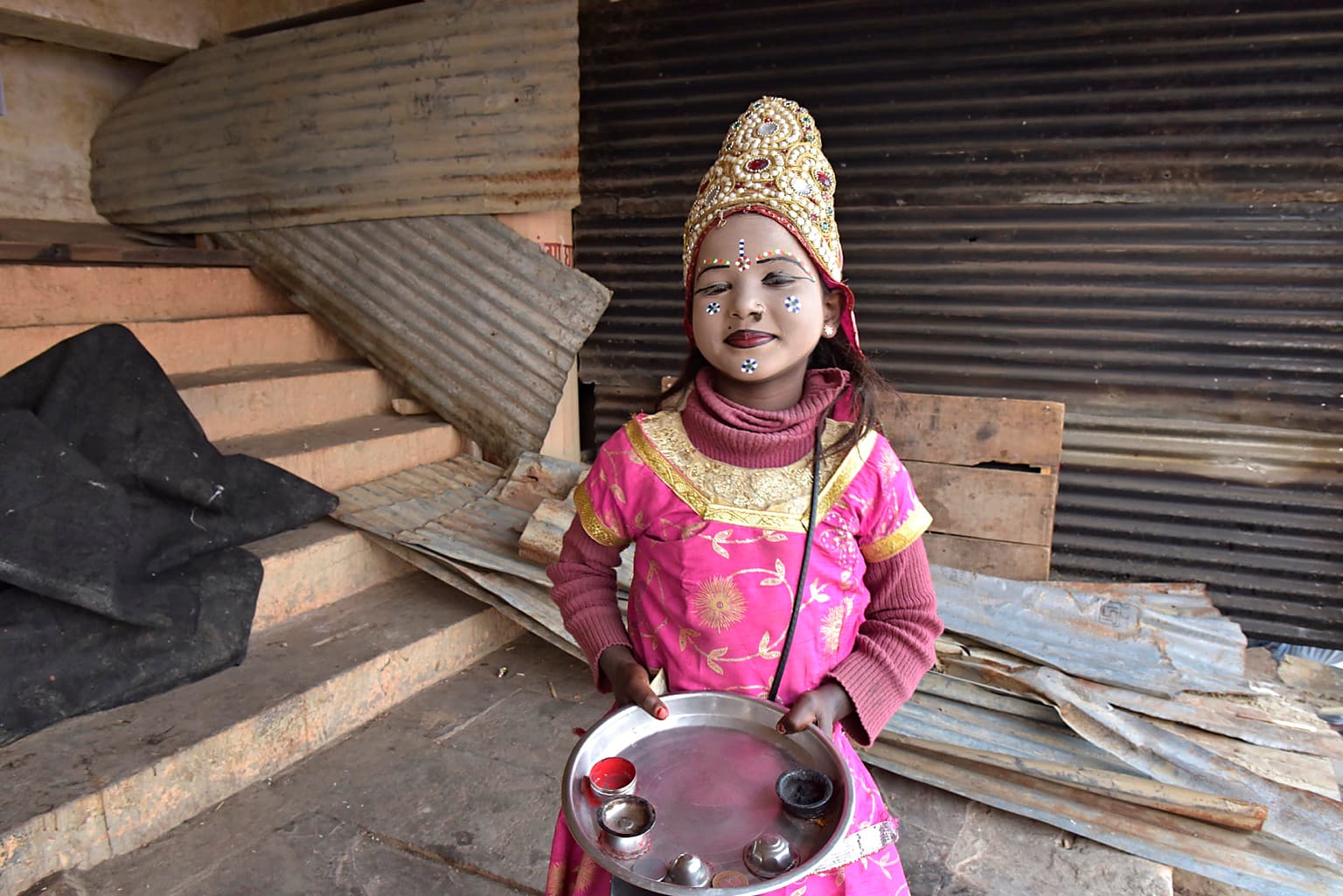 A young girl in bright pink ceremonial dress stands before corrugated shutters holding a metal tray in Varanasi, 2019