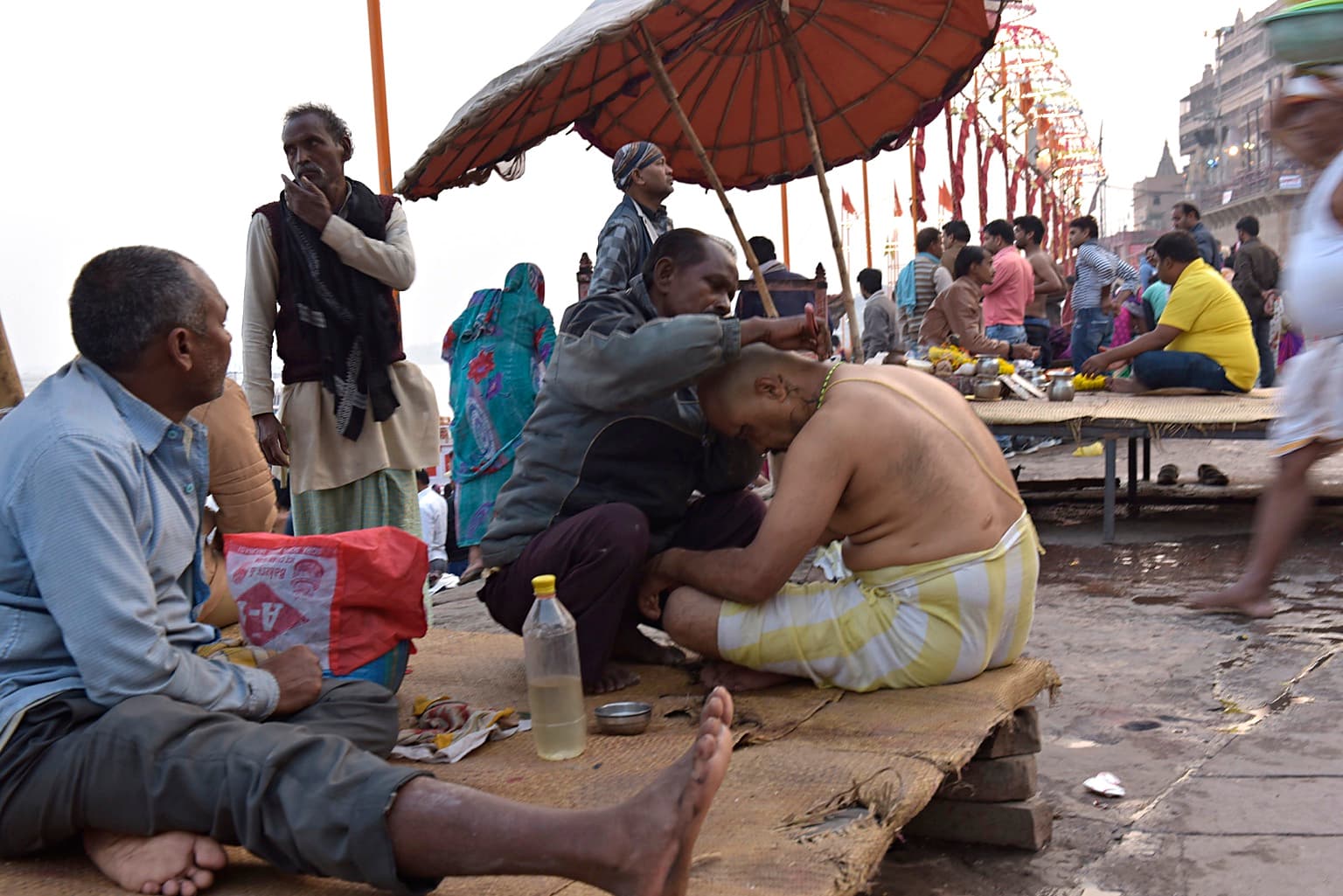 A man works on another man’s hair during a crowded riverside gathering in Varanasi, 2019