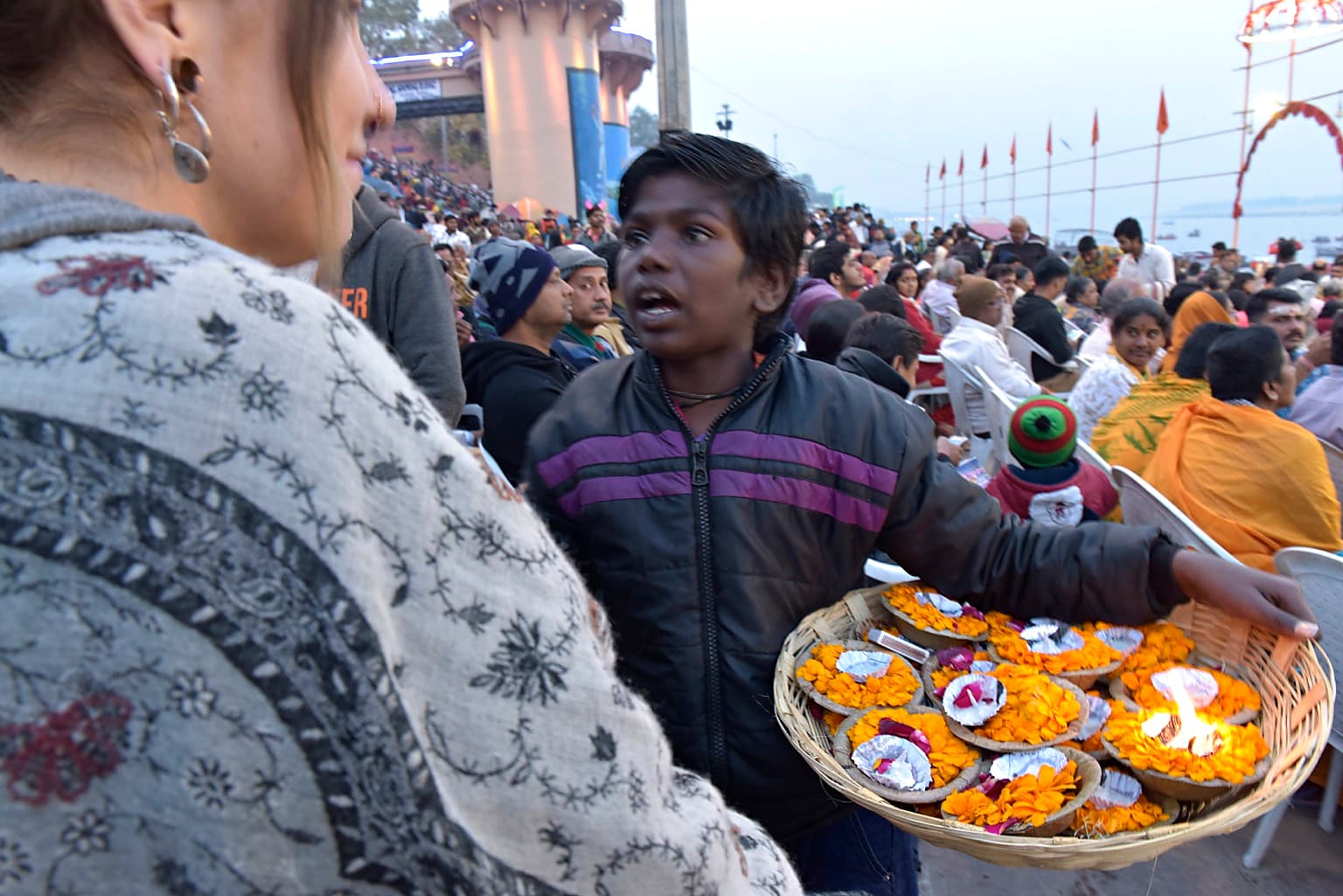 A young vendor carrying a broad basket of marigold offerings speaks with a visitor in a crowded gathering in Varanasi, 2019