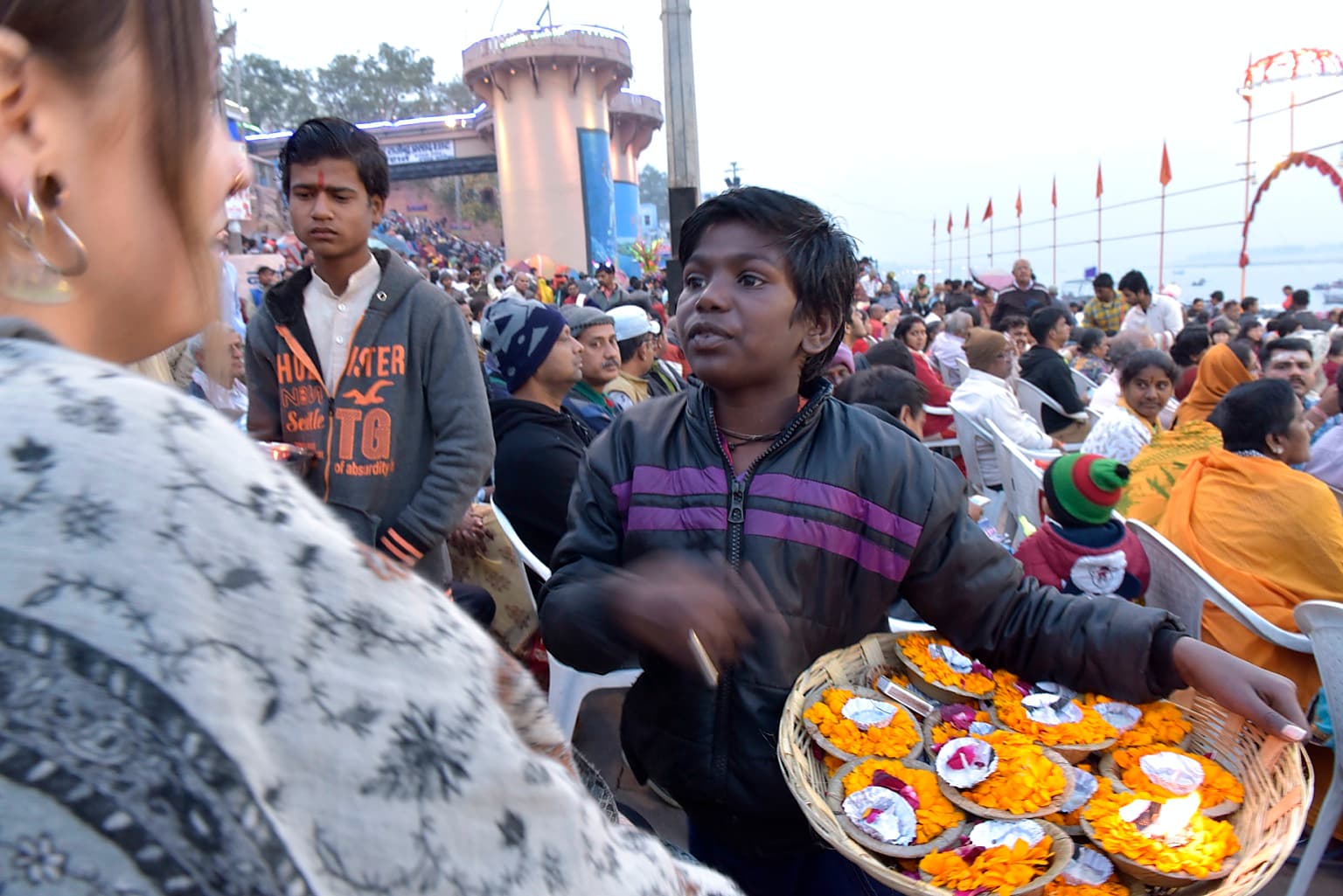 A young vendor gestures while speaking with a visitor beside a basket filled with marigold offerings in Varanasi, 2019