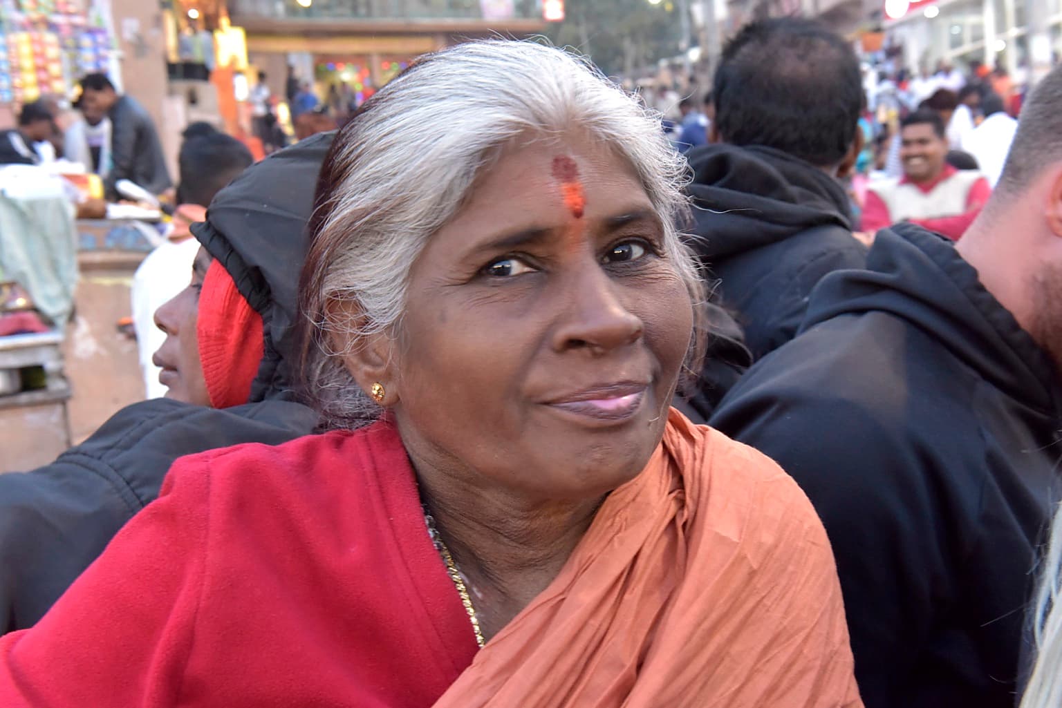 An older woman with silver hair and red and ochre cloth looks directly toward the camera in Varanasi, 2019