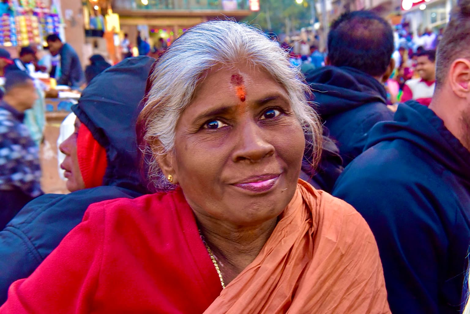 An older woman with silver hair and a vivid red wrap meets the camera in a crowded Varanasi gathering, 2019