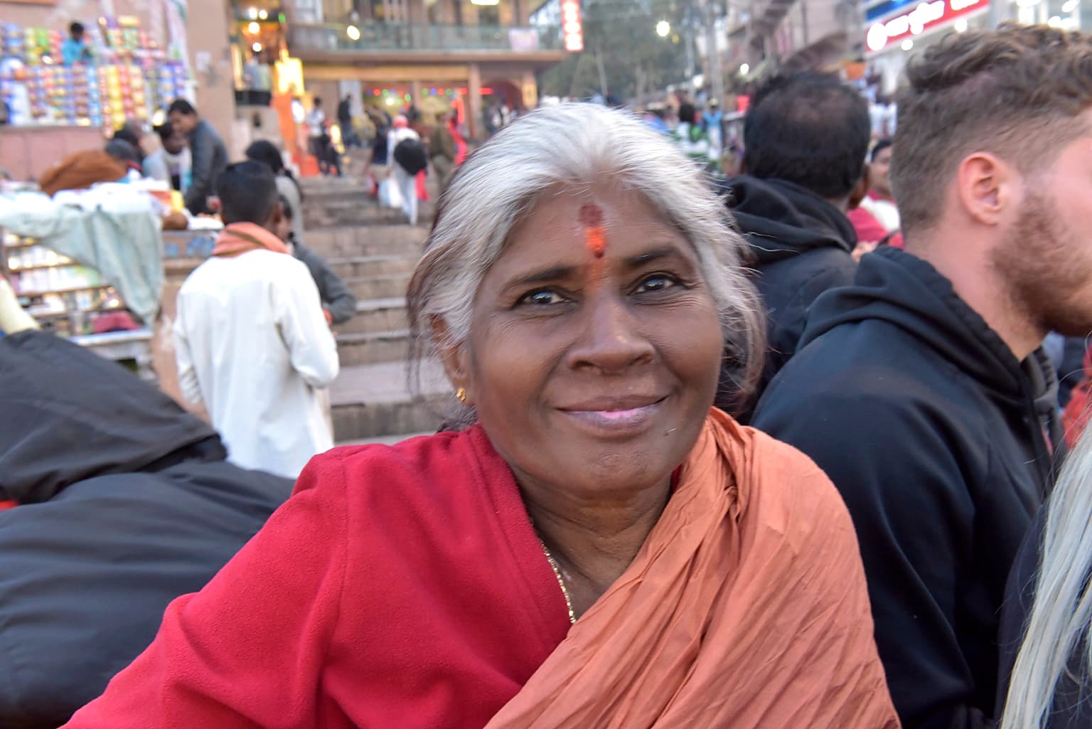 An older woman with silver hair smiles toward the camera amid a crowded gathering in Varanasi, 2019