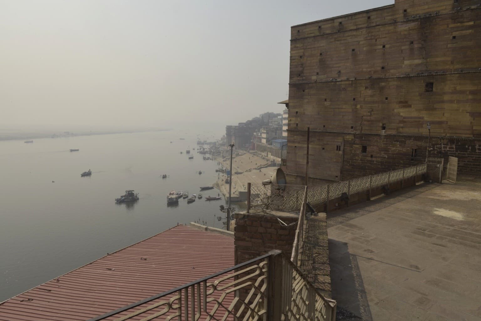 A high terrace view looks past the massive wall of a riverfront mosque toward boats and distant ghats along the Ganges in Varanasi, 2019