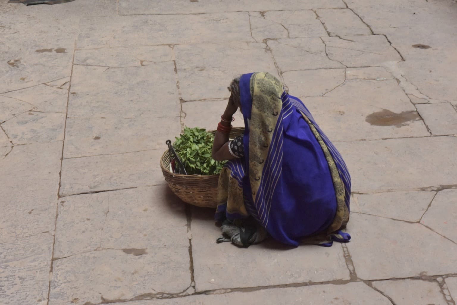 A woman in a deep blue sari sits beside a large woven basket of leafy greens on stone paving in Varanasi, 2019