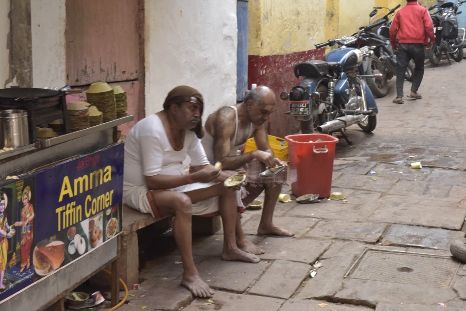 Two men sit beside an Amma Tiffin Corner stall eating in a narrow Varanasi lane, with motorcycles parked behind them, 2019