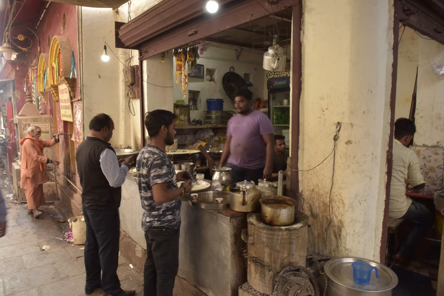 Customers stand at a compact food stall in a narrow Varanasi lane while a saffron-robed man passes at left, 2019