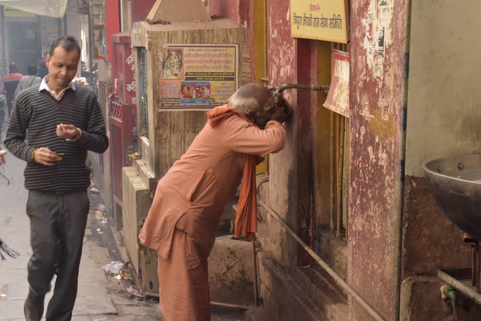 A saffron-robed man leans to drink from a wall-mounted water spout in a narrow Varanasi lane, 2019