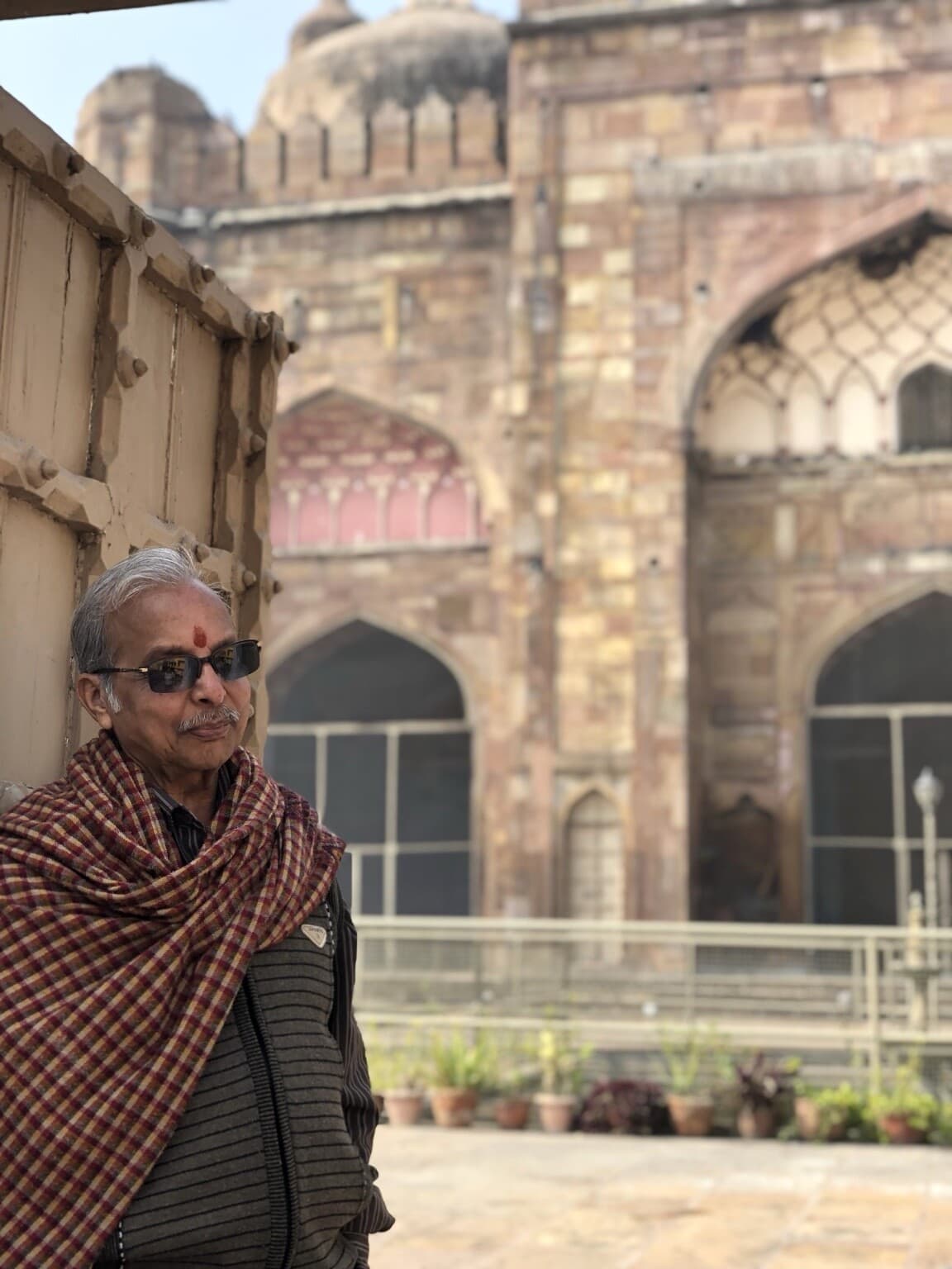 An older man wearing sunglasses and a checked shawl stands beside the mosque courtyard in Varanasi, 2019