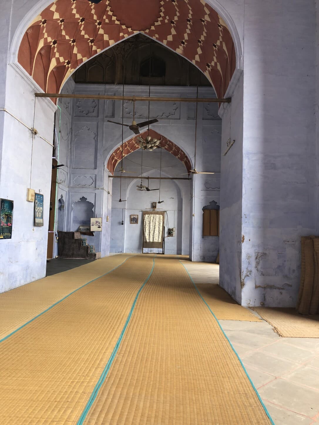 An empty central aisle leads through a blue-walled mosque interior beneath repeating arches and hanging fans in Varanasi, 2019