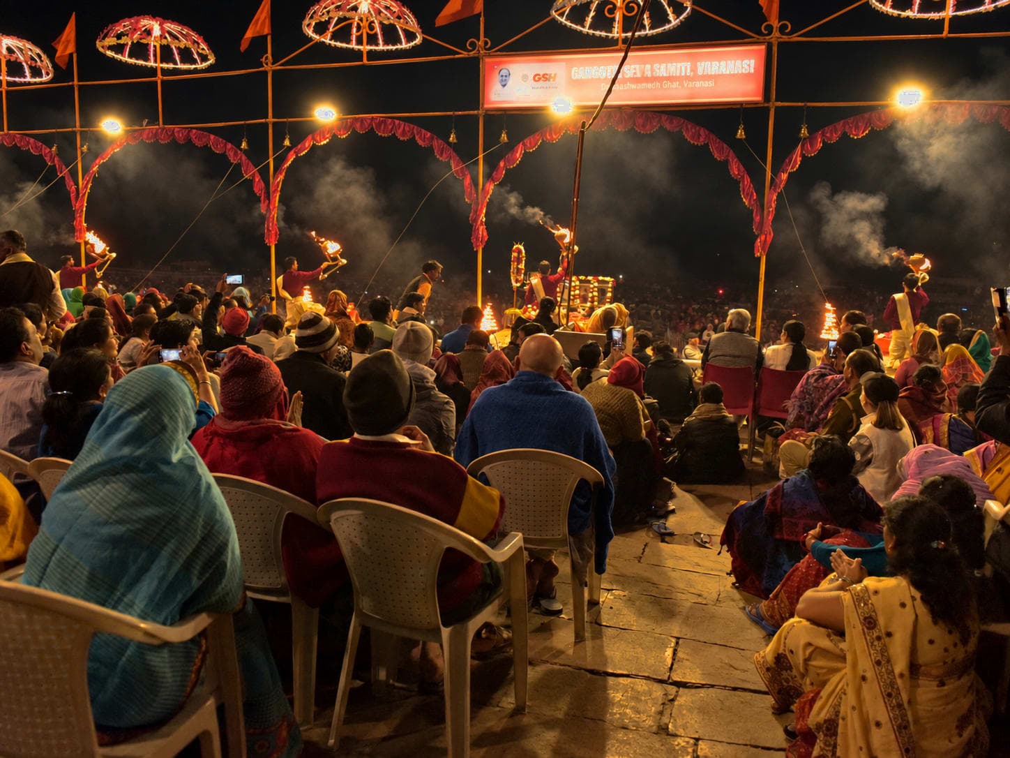 A broad view of evening Ganga aarti shows multiple priests, flames and a large seated crowd beneath repeating arches in Varanasi, 2019