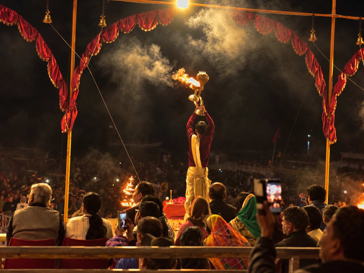 A priest seen from behind raises a smoking ritual vessel beneath an illuminated arch during evening Ganga aarti in Varanasi, 2019